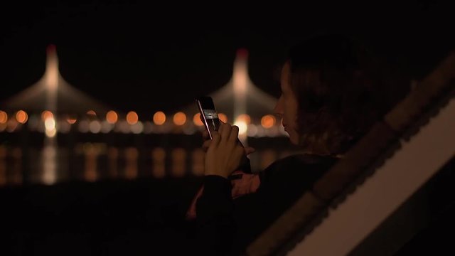 Young attractive lady sitting in the night park and wathing social media feed on her brand new mobile phone.
