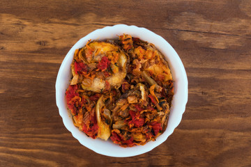Vegetable appetizer of fried zucchini rings with tomatoes, onions and carrots in a white porcelain bowl on a wooden background, top view.