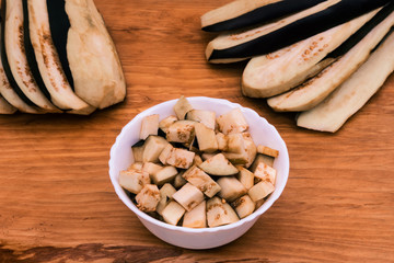 A white bowl filled with sliced eggplant on a wooden table next to slices of eggplant.