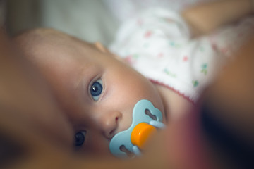 Close-up portrait of cute little baby girl with blue pacifier is lying on the bed in bedroom near...