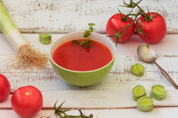 Clear red tomato soup in the green bowl among vegetables on the old white shabby planks