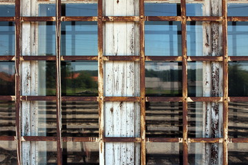 Rusted metal bars in shape of net mounted on old dilapidated white wooden window frame with windows reflecting houses, trees and clear blue sky in background