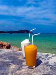 Two fruit shakes at the beach, with blue sky, calm ocean and islands in the background and the drinks and a concrete table in the foreground. Taken in Chaloklum, Ko Pha Ngan, Thailand.