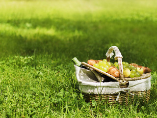 Wicker basket for picnic with green grape, apples, bottle of wine and croissants standing on green grass
