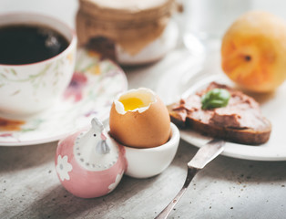 Egg soft-boiled with a salt-cellar in the form of chicken. A cup of coffee, a toast of black bread with pate, a peach - all in the background.