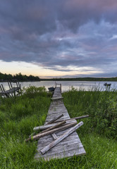 Rustic wooden pier with broken posts leading into lake under sunset sky