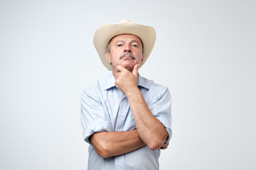 Mature man adjusting his cowboy hat and looking at camera while standing against grey background