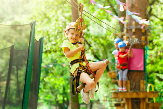 Brave young girl in helmet climbs on tree tops in amusement rope park on summer holidays, children camp