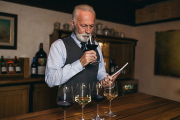Mature sommelier smelling flavor of red wine in glass, he is checking color, quality and sediments of drink.