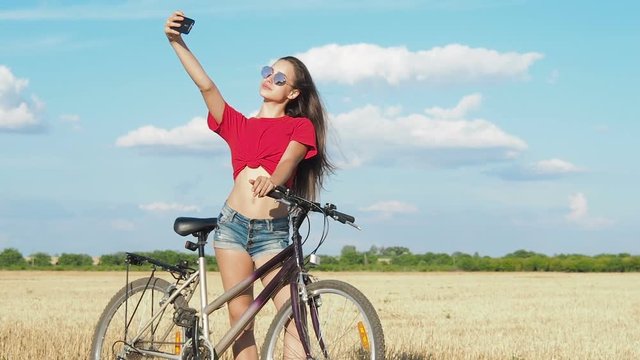 The girl makes selfie. A beautiful girl with a bicycle takes a picture of herself on the phone.
