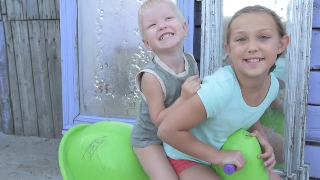 Children ride on green plastic horse rocking on background of mirrors in courtyard of rural house. Beautiful kids, Brother and sister are happy together on vacation in village.