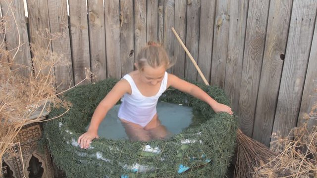 Young girl is preparing to take part in celebration of Halloween. Child is bathing in cauldron with chamomile flowers, trying on pointed hat. She is ready to fly on broomstick like a witch.