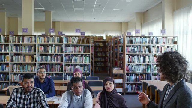 Tilt down of female teacher standing at flipchart in classroom and explaining something to group of adult migrants while giving free language course