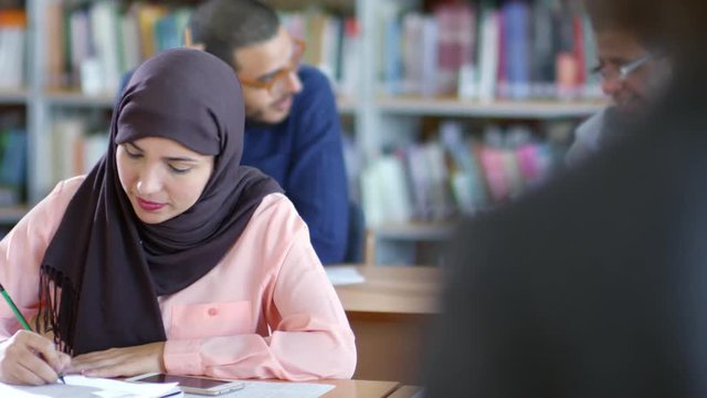 Tilt down of attractive muslim woman in hijab sitting at desk, writing in notebook and listening to teacher at college