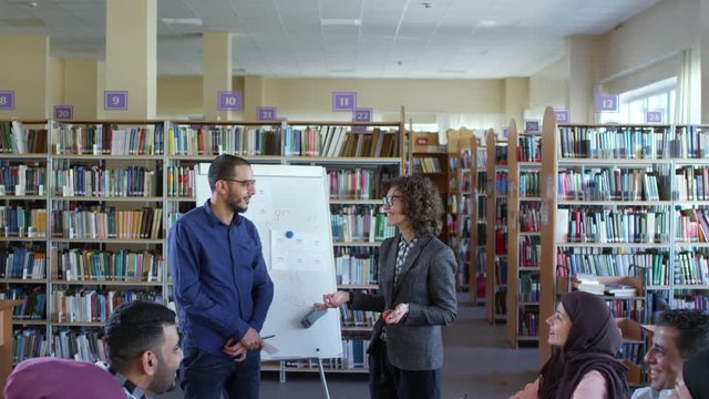 Tilt down of young middle eastern man standing by flipchart in classroom and showing word card to Caucasian female teacher and group of migrant students at language lesson