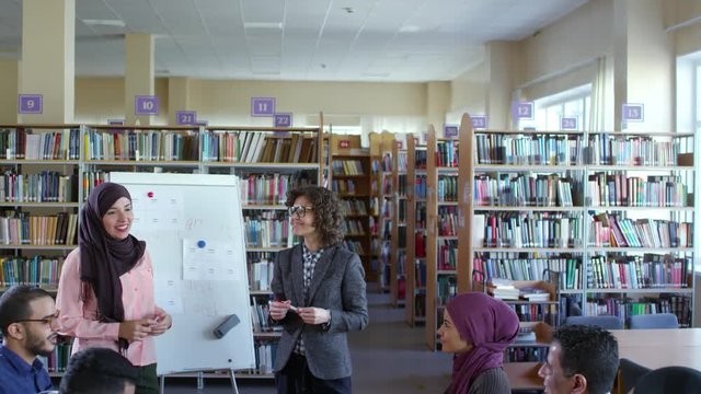 Tilt down of muslim woman in hijab standing by flipchart in classroom, smiling and talking with Caucasian female teacher and group of migrant students at language lesson