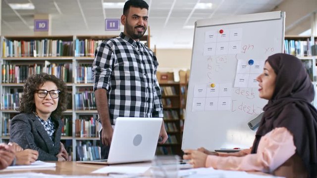 Cheerful middle eastern man standing by flipchart at English lesson for migrants at talking with group of adult students and female teacher