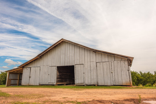 An old, beautiful barn that holds farm equipment stands regally on the side of the road in rural, upstate South Carolina. Bright blue sky and fluffy clouds behind.