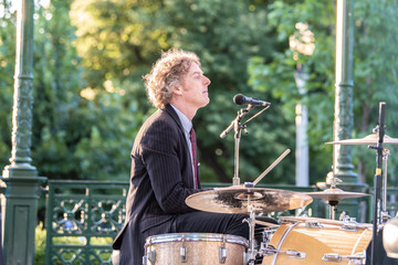 Attractive forty something man plays drums in a band, outside in a park, on a gazebo stage.