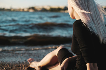 Fototapeta na wymiar Young blonde woman sitting on the shore and looking at the water, sunset light