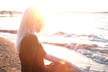 Young blonde woman sitting on the shore and looking at the water, sunset light