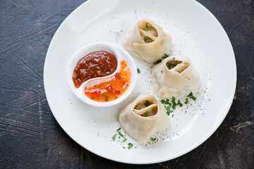Steamed meat dumplings manti with sauces and greens served on a white plate, elevated view over dark brown stone background