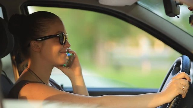 young beautiful woman with red hair in sunglasses sitting behind the wheel of a car with a phone in her hand at sunset on a summer day.