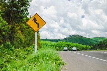 Warning reverse curve left traffic sign with vehicles on road. Landscape view of roadside at countryside in Thailand.