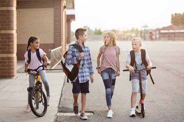 Diverse group of school kids talking and walking home from school together. Full length candid photo of both boys and girls leaving the school building on their bikes and scooters - Powered by Adobe