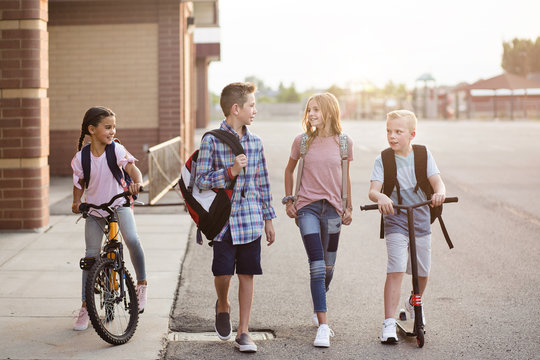 Diverse group of school kids talking and walking home from school together. Full length candid photo of both boys and girls leaving the school building on their bikes and scooters