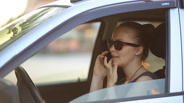 young beautiful woman with red hair in sunglasses sitting behind the wheel of a car with a phone in her hand at sunset on a summer day.