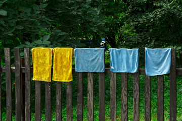 Multicolored rags hang and dry on a fence of damp wooden laths against the background of green leaves. Rags hang on the fence. Multicolored old towels for washing the floor are dried on the fence.