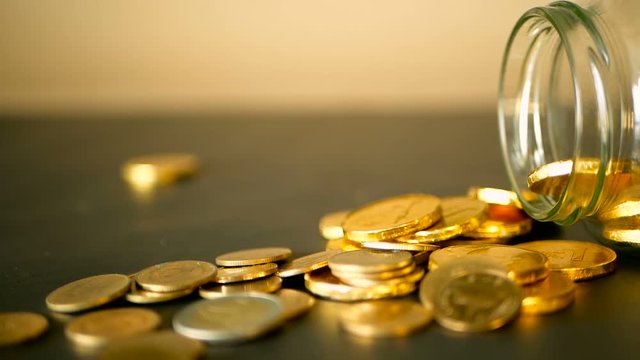 Yellow coins fell out from jar. Symbol of investing, keeping money concept. Collecting cash conis in glass tin as moneybox. Close-up still life with gold coins on black table and rotating penny.