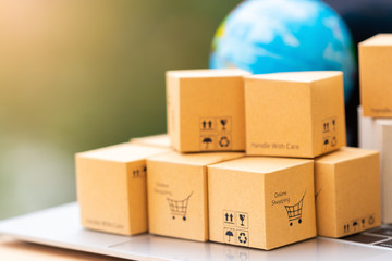 Fototapeta premium Stack of cardboard boxes with warning and shopping cart icon on laptop keyboard with globe nearby, with nature green background. Consumer buy product anywhere. Online shopping and e-commerce concept.