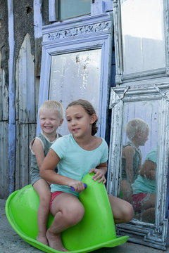 Children ride on green plastic horse rocking on background of mirrors in courtyard of rural house. Beautiful kids, Brother and sister are happy together on vacation in village.