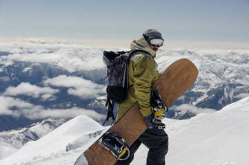 man in ski equipment, wearing safety glasses, rises to a snowy mountain against a cloudy sky - obrazy, fototapety, plakaty