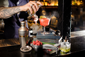 Barman spraying peat bitter on a glass with red sweet summer cocktail arranged on books