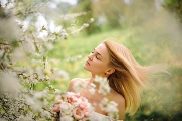 Fototapeta premium Tender young blonde woman in white dress with a bouquet near the blooming cherry tree with closed eyes