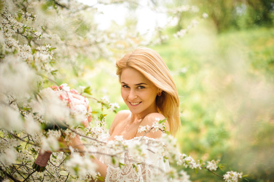 Smiling young blonde woman in white dress with a bouquet near the blooming cherry tree