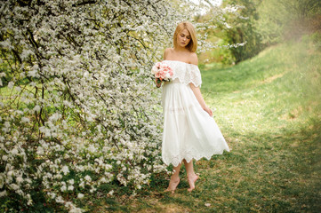 Romantic full height portrait od a blonde woman in white dress with a bouquet standing near the blooming tree