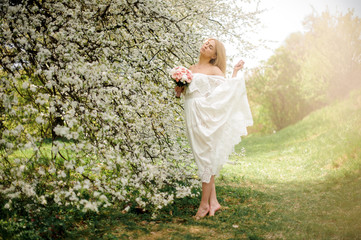 Romantic full height portrait od a blonde woman in white dress with a bouquet standing on tiptoe...