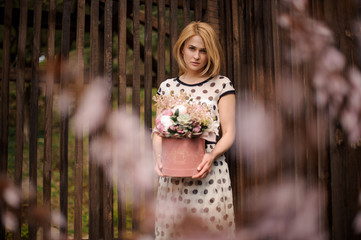 Blonde woman holding a flower box standing on the background of wooden fence and pink blooming tree
