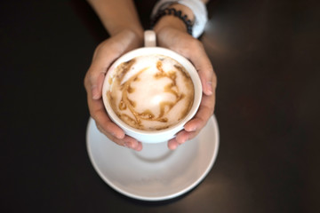 White cup of caramel coffee on woman's hands for charge energy, refresh, breakfast or breaking time in afternoon. Cappuccino hot coffee on top with foam milk and caramel cream.