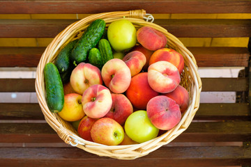 Woven from a vine basket stands on wooden boards filled with fruits from a fresh harvest. Peaches, apples, cucumbers.