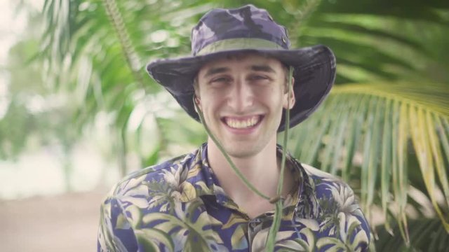 Portrait of Young Male explorer in tropical beach setting. Smiling into the camera as he adjusts his wide brim hat