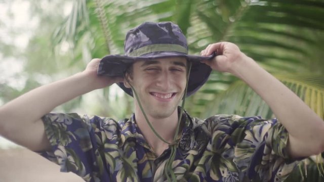 Attractive young white male in explorer outfit puts on his wide brim hat as he smiles into the camera. Shot in tropical setting.