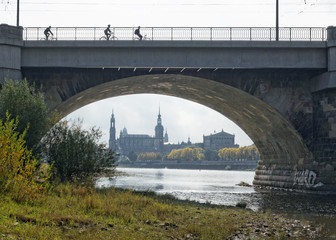 Dresden - view through an arch of the Marienbrücke to the city with Hofkirche, castle and Semperoper, low tide of the Elbe, autumnal colored, backlit with a slight haze, silhouettes of cyclists