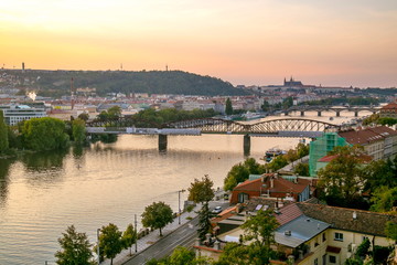 Fototapeta premium View from Vysehrad of Prague castle, sunset sky with pink and yellow colors, railway bridge, stone bridge, river Vltava, boats, riverbank, streets, cars, houses, red roofs, green trees, hill of Petrin