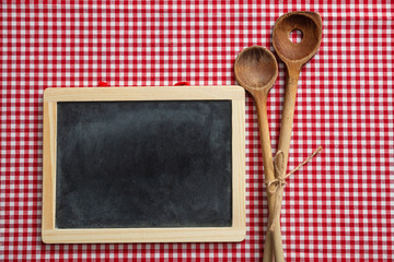 Blackboard and wooden kitchen utensils on red checkered picnic tablecloth, space for text