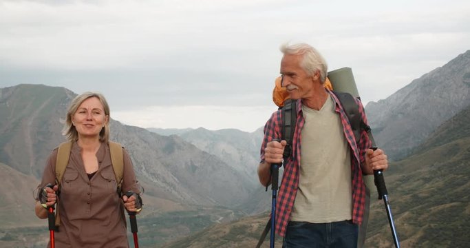 Positive mature caucasian couple trekking in mountains with backpacks, enjoying their adventure 4k